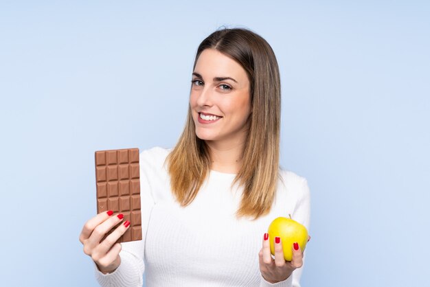 Jovem mulher loira sobre parede azul isolada, tomando uma tablete de chocolate em uma mão e uma maçã na outra