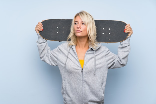 Jovem mulher loira sobre parede isolada com skate e olhando de lado