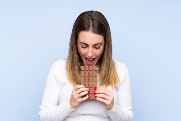 Jovem mulher loira sobre parede isolada, comendo uma tablete de chocolate