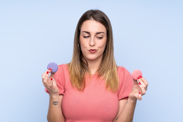 Jovem mulher loira sobre parede isolada segurando macarons franceses coloridos e infeliz