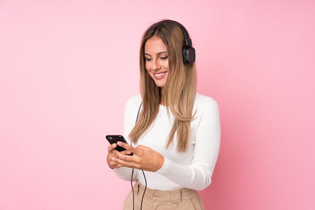 Jovem mulher loira sobre rosa isolado usando o celular com fones de ouvido