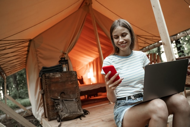 Jovem mulher loira sorridente, sentada na área de glamping usando laptop e mensagens no smartphone. Estilo de vida de acampamento. Viagem de baixo orçamento. Conceito de trabalho remoto. Tecnologia de comunicação de internet moderna.