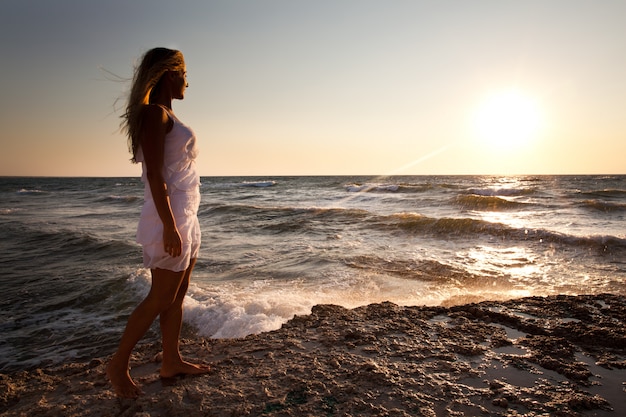 Jovem mulher loira sorridente vestido branco de pé nas rochas e olhando o pôr do sol sobre a água do mar ondulada