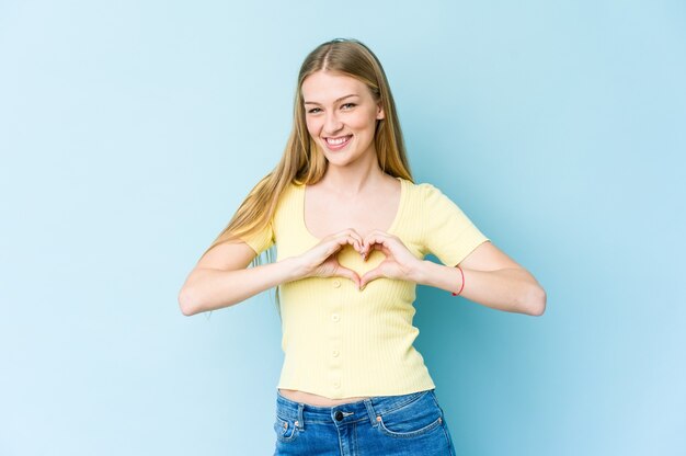 Jovem mulher loira sorrindo e mostrando uma forma de coração com as mãos.