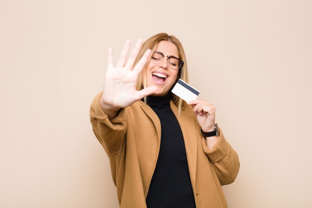 Jovem mulher loira sorrindo e olhando amigável, mostrando o número cinco ou quinto com a mão para a frente, contando com um cartão de crédito