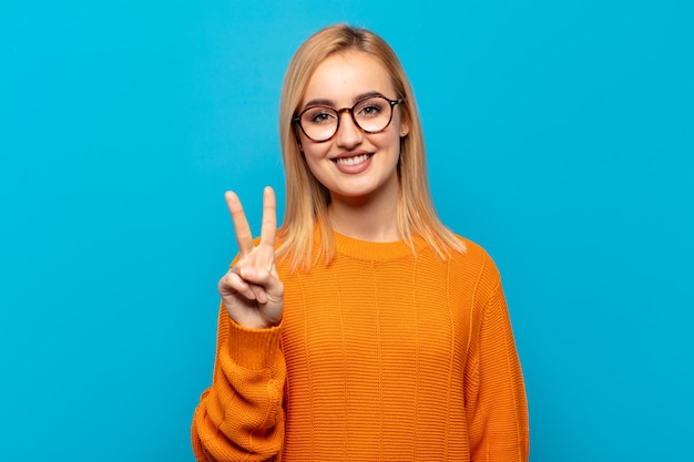 Jovem mulher loira sorrindo e parecendo amigável, mostrando o número dois ou o segundo com a mão para a frente