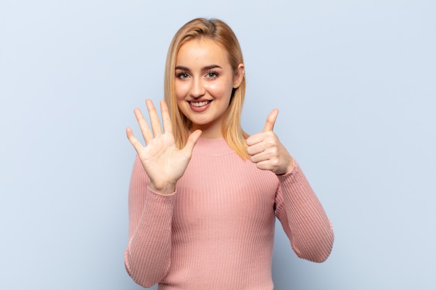 Jovem mulher loira sorrindo e parecendo amigável, mostrando o número seis ou sexto com a mão para a frente, em contagem regressiva