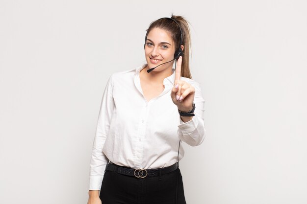Jovem mulher loira sorrindo e parecendo amigável, mostrando o número um ou primeiro com a mão para a frente, em contagem regressiva