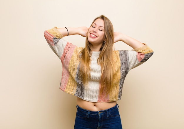 Jovem mulher loira sorrindo e se sentindo relaxado, satisfeito e despreocupado, rindo positivamente e relaxando contra a parede bege