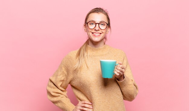 Jovem mulher loira sorrindo feliz com uma mão no quadril e uma atitude confiante, positiva, orgulhosa e amigável. conceito de café