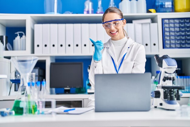 Jovem mulher loira trabalhando no laboratório cientista apontando o polegar para o lado sorrindo feliz com a boca aberta