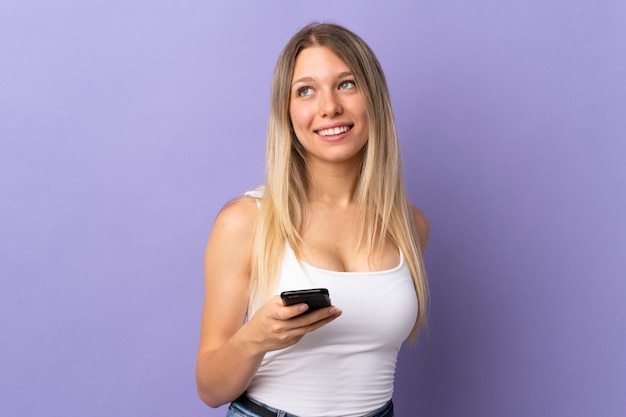 Jovem mulher loira usando celular isolada em roxo rindo e olhando para cima