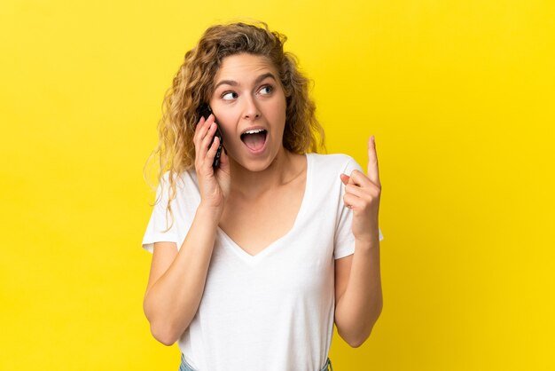 Jovem mulher loira usando celular isolado em um fundo amarelo pensando em uma ideia apontando o dedo para cima