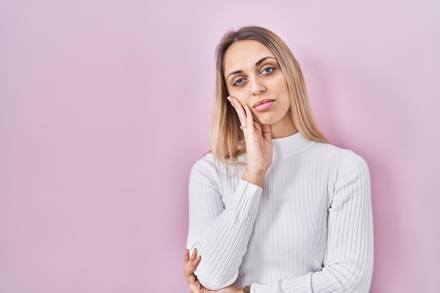 Jovem mulher loira vestindo suéter branco sobre fundo rosa pensando parecendo cansada e entediada com problemas de depressão com braços cruzados