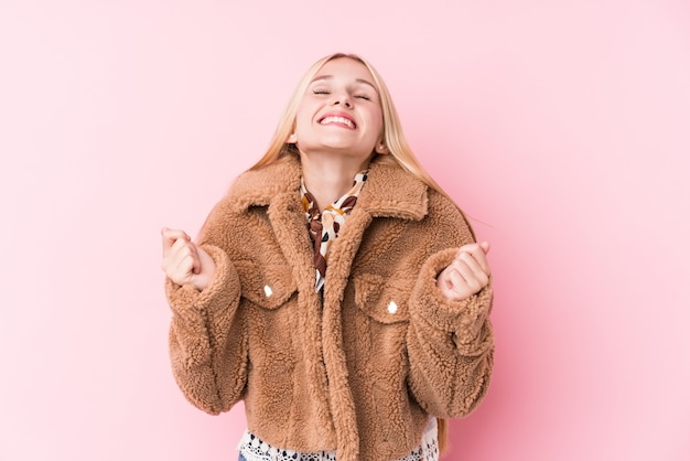 Jovem mulher loira, vestindo um casaco contra uma parede rosa, levantando o punho, sentindo-se feliz e bem sucedido. Conceito de vitória