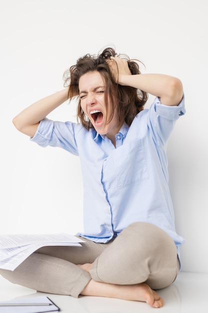 Foto jovem mulher louca e estressada em trajes casuais sentada isolada, despenteando o cabelo e gritando enquanto expressa o fracasso