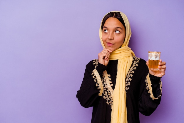 Jovem mulher marroquina segurando um copo de chá isolado no fundo roxo, olhando de soslaio com expressão duvidosa e cética.