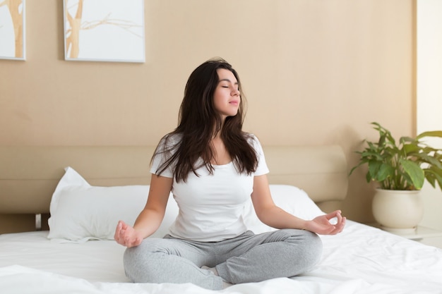 Jovem mulher meditando e relaxando enquanto está sentada na cama em casa