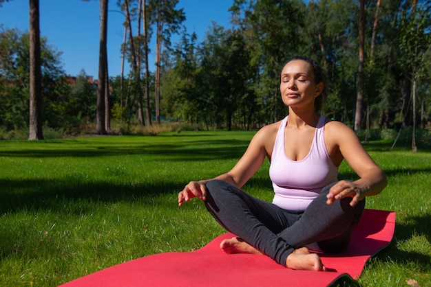 Jovem mulher meditando enquanto está sentada em uma esteira de ioga na grama verde no belo parque de pinheiros