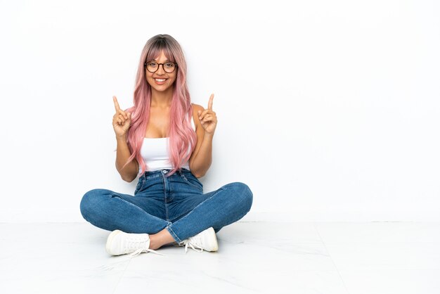 Jovem mulher mestiça de cabelo rosa sentada no chão isolada no fundo branco apontando uma ótima ideia