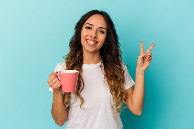 Jovem mulher mexicana segurando uma caneca isolada sobre fundo azul alegre e despreocupada, mostrando um símbolo de paz com os dedos.