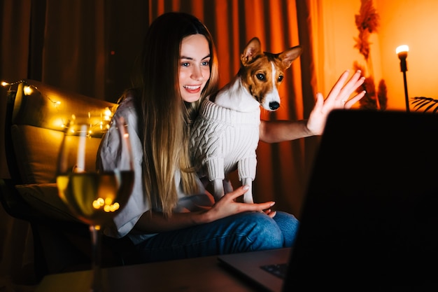 Foto jovem mulher milenar com seu cachorro, tendo uma videochamada no laptop e bebendo vinho, usa a tecnologia para se comunicar com amigos ou família.