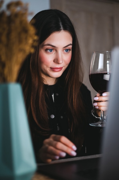 Jovem mulher milenar fazendo videochamada no laptop e bebendo vinho, usa a tecnologia para se comunicar com amigos ou familiares