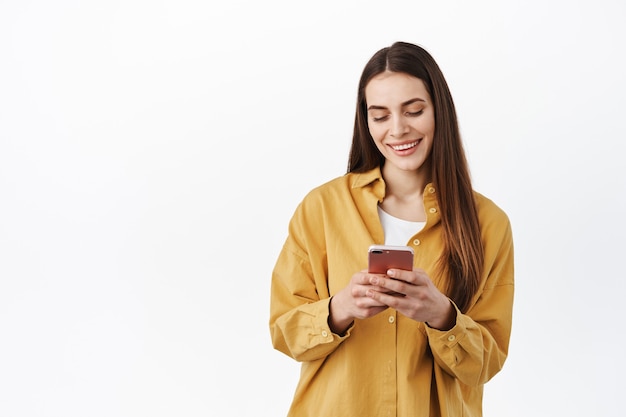 Jovem mulher moderna conversando ao telefone, lendo a tela do smartphone com um sorriso despreocupado, enviando mensagens de texto ou navegando no aplicativo da loja online, encostada na parede branca