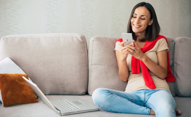 Jovem mulher moderna e confiante e sorridente está trabalhando ou relaxando em casa e sentada no sofá enquanto usa seu smartphone
