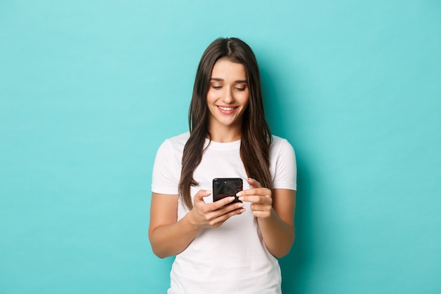 Jovem mulher moderna em t-shirt branca, usando telefone celular. Menina trocando mensagens e sorrindo, em pé