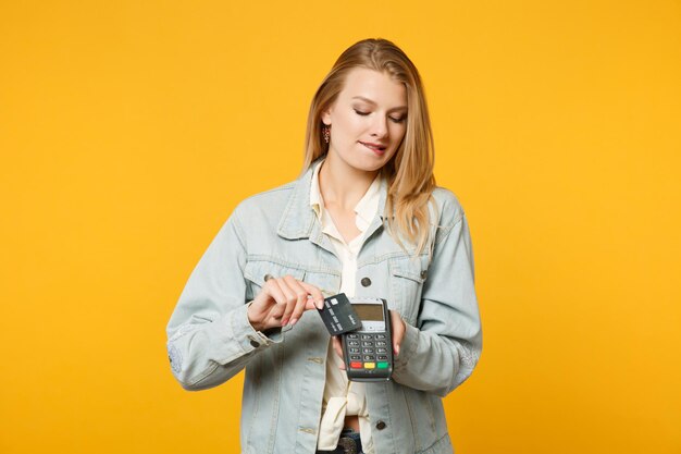 Jovem mulher mordendo os lábios, segurando o terminal de pagamento bancário moderno sem fio para processar e adquirir pagamentos com cartão de crédito isolados em fundo amarelo laranja. Conceito de estilo de vida de pessoas. Mock up espaço de cópia.