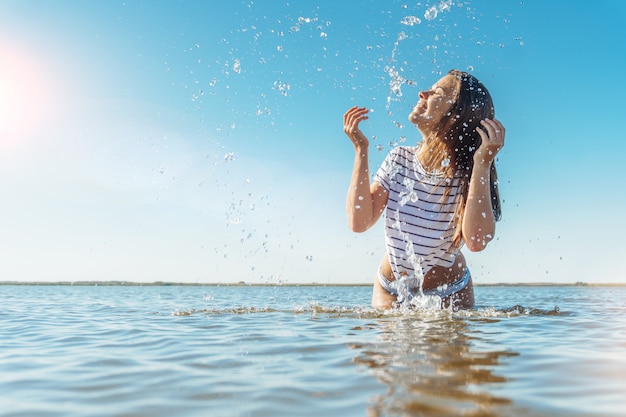 Jovem mulher morena atraente na camisa listrada branca e fio dental, salpicos de água em um mar com céu azul