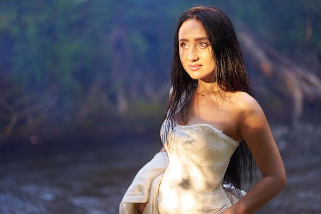 Jovem mulher morena bonita no vestido de casamento branco ao ar livre perto do rio com os cabelos molhados na noite a luz do sol no rosto