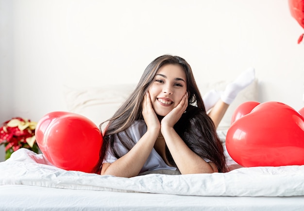 Jovem mulher morena feliz deitada na cama com balões em forma de coração vermelho