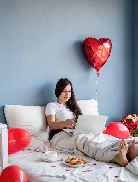 Jovem mulher morena feliz sentada na cama com balões em forma de coração vermelho trabalhando no laptop