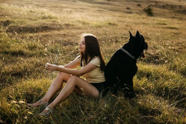 Jovem mulher morena sentada em um campo ao lado de um grande cão preto de raça schnauzer gigante