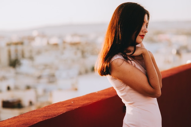 Jovem mulher moreno bonita em um vestido de noite branco. cenário pôr do sol cenário da cidade.