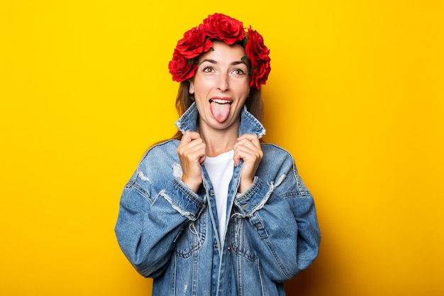 Jovem mulher mostrando a língua na jaqueta jeans e coroa de flores vermelhas na cabeça na parede amarela.
