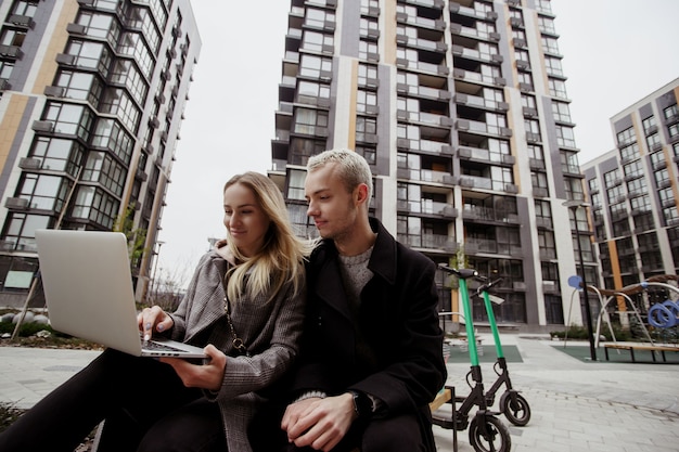 Foto jovem mulher mostrando fotos para seu amigo masculino e sorrindo. homem loiro sentado perto e olhando no laptop. eles se sentam em bancos perto de blocos de apartamentos. passando um bom tempo. duas e-scooters próximas.