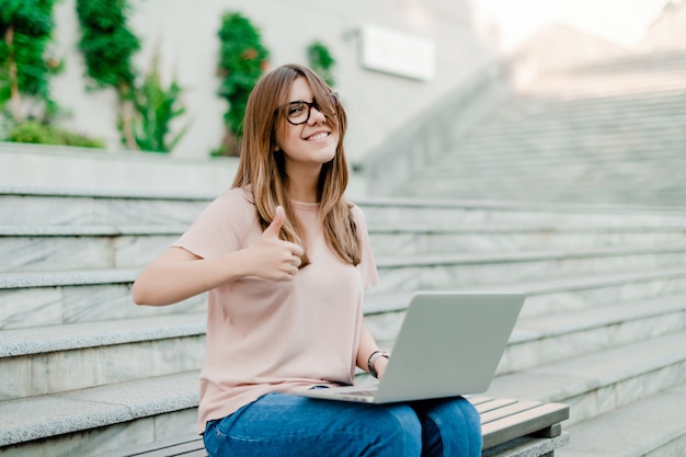 Jovem mulher mostrando os polegares e usando o laptop para trabalhar freelance ao ar livre