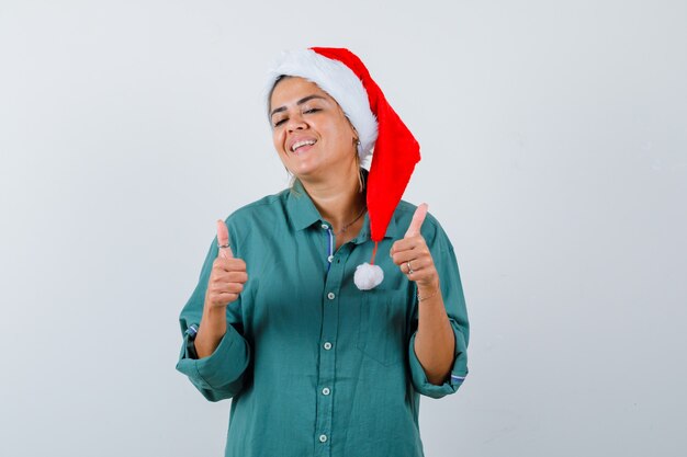 Jovem mulher mostrando os polegares na camisa, chapéu de Papai Noel e olhando satisfeita, vista frontal.