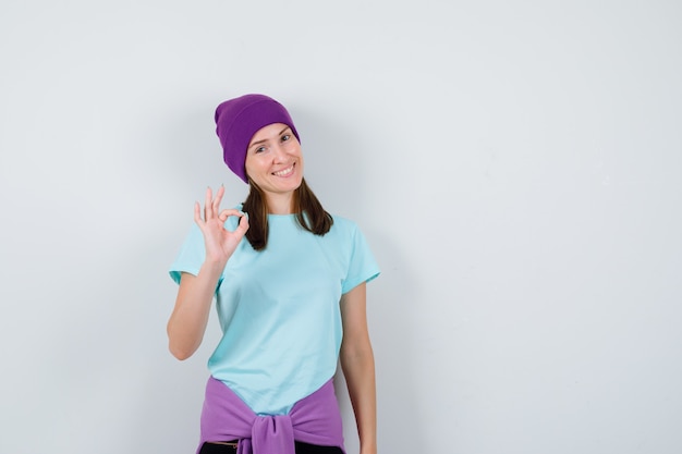 Jovem mulher mostrando sinal de tudo bem em t-shirt azul, gorro roxo e olhando alegre, vista frontal.