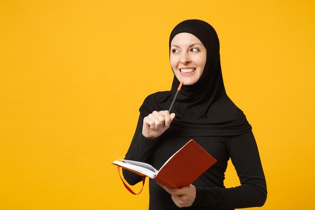 Jovem mulher muçulmana árabe em roupas pretas de hijab segurar o caderno, escrever, preparar-se para o exame isolado no retrato de parede amarela. conceito de estilo de vida religioso de pessoas. .