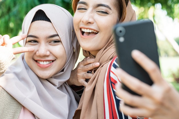 Jovem mulher muçulmana asiática com lenço na cabeça encontrar amigos e usar o telefone no parque para uma selfie ou videochamada