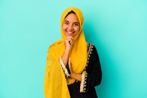 Jovem mulher muçulmana isolada em um fundo azul, sorrindo feliz e confiante, tocando o queixo com a mão.