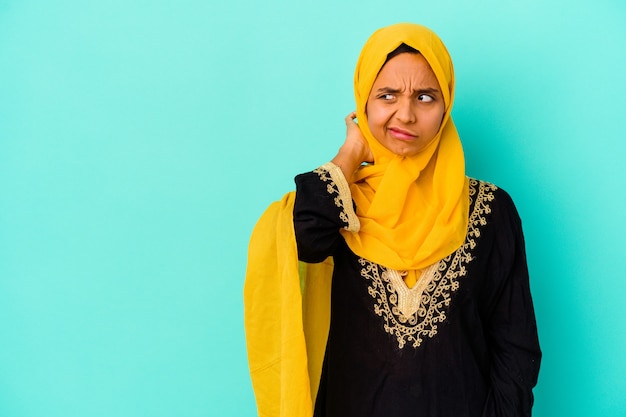 Jovem mulher muçulmana isolada na parede azul, tocando a parte de trás da cabeça, pensando e fazendo uma escolha.