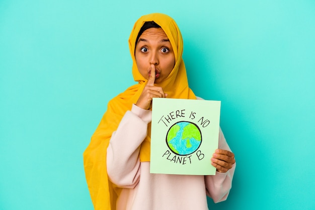 Jovem mulher muçulmana segurando um cartaz de não há planeta b isolado na parede azul, mantendo um segredo ou pedindo silêncio