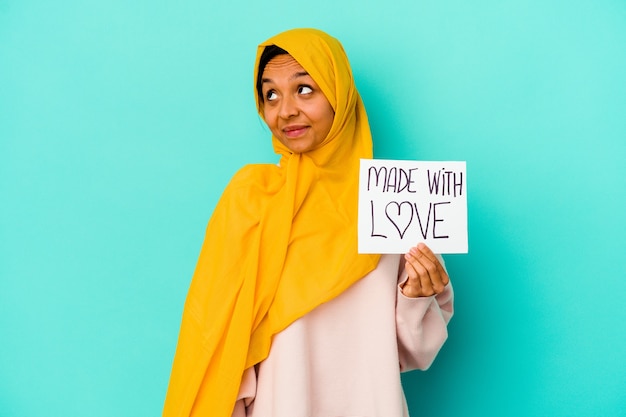 Jovem mulher muçulmana segurando um cartaz feito com amor isolado na parede azul, sonhando em alcançar objetivos e propósitos