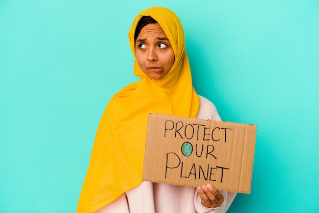 Jovem mulher muçulmana segurando um proteger nosso planeta isolado em um fundo azul confuso, sente-se em dúvida e inseguro.