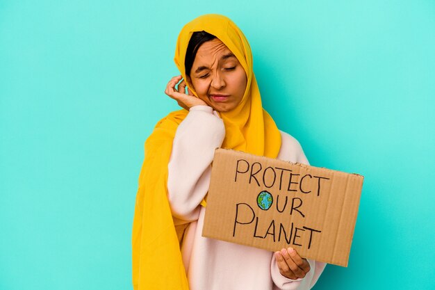Jovem mulher muçulmana segurando um proteger nosso planeta isolado na parede azul, cobrindo as orelhas com as mãos.